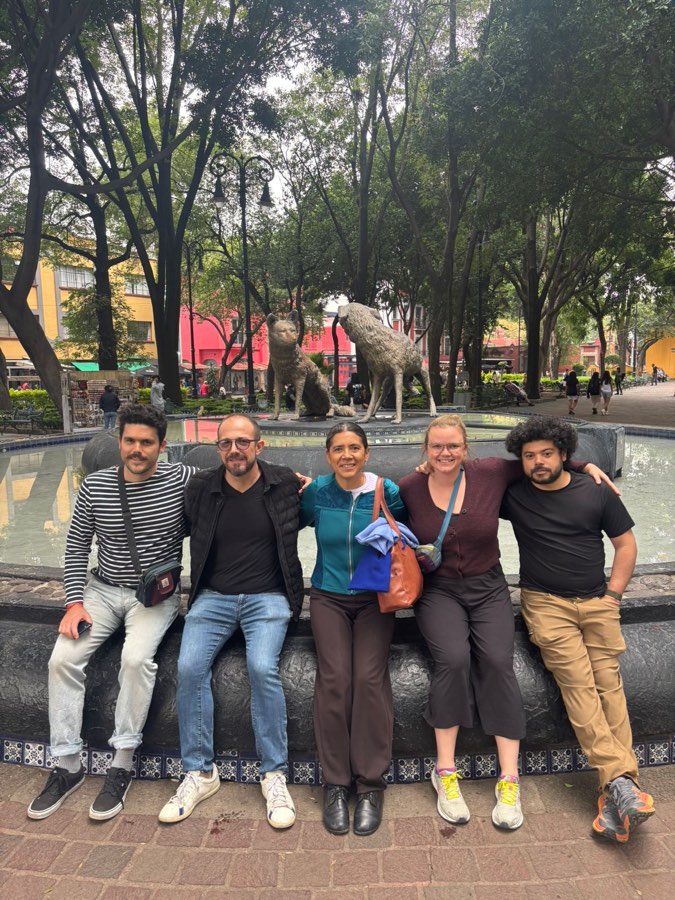 Five travelers sitting together on the edge of a fountain in a tree-lined plaza